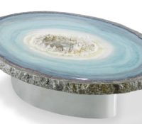 Glacier Geode Table<br />
A cool-toned inspired agate encased and molded into a stone frame. Resin, in various shades of blue and natural hues poured in layers over an acrylic base. Creating large ring patterns receding into a pool of natural Brazilian clear quartz point structures. Mirroring the shape, a custom steel base holds this beautiful 3-inch-thick oval slab.<br />
